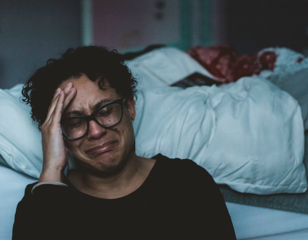 An African American woman wearing glasses crying, sitting on the floor next to white bed.