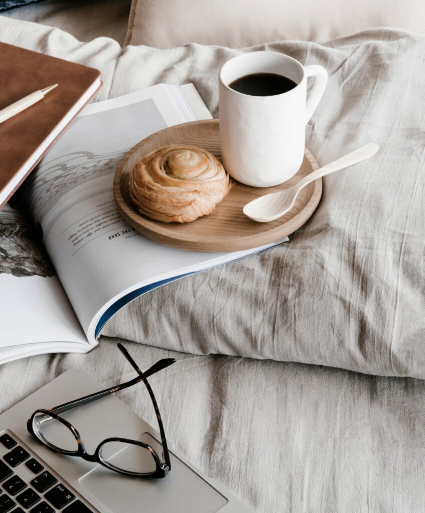 A cup of coffee with donut on pillow with paperwork, glasses and laptop.