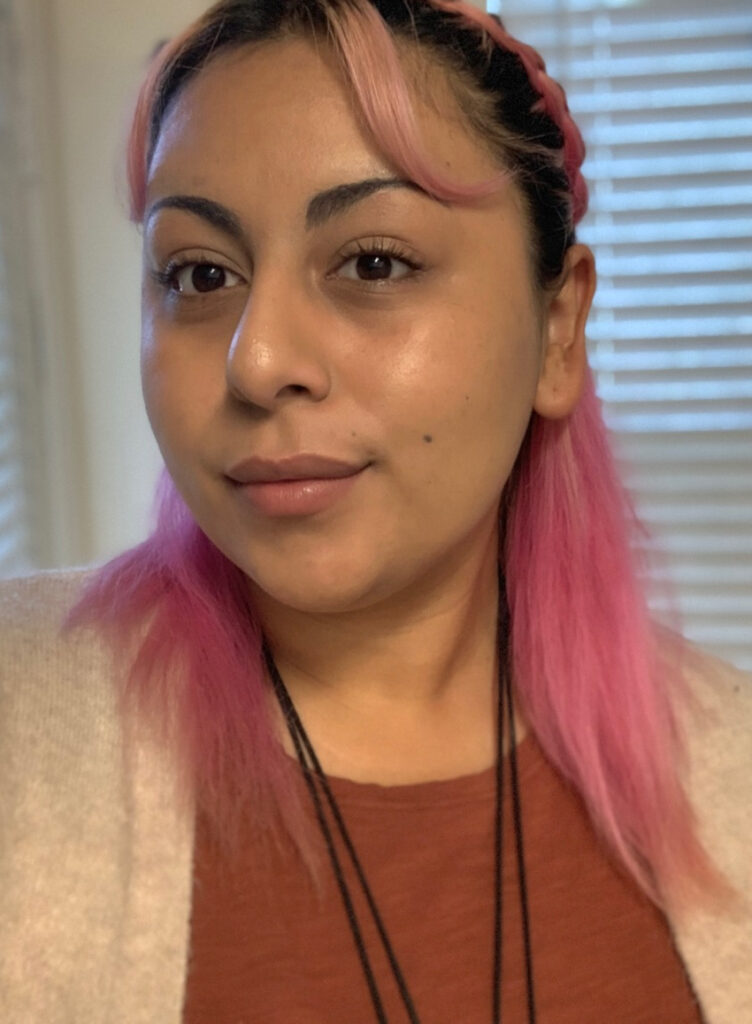 A young woman with pink hair wearing a tan sweater over a brown shirt.