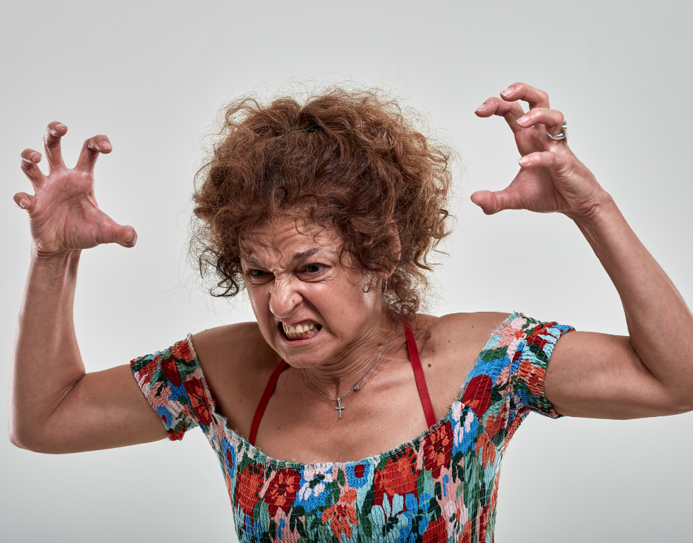 An angry woman with reddish-brown hair and wearing a colorful top with her hands gnarled in the air.