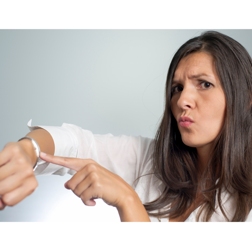 A white woman wearing a white shirt while frowning and pointing at her watch.